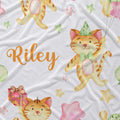 A white fabric blanket with a whimsical watercolor print of orange tabby cats, pastel stars, balloons, and the name 'Riley' in orange script.