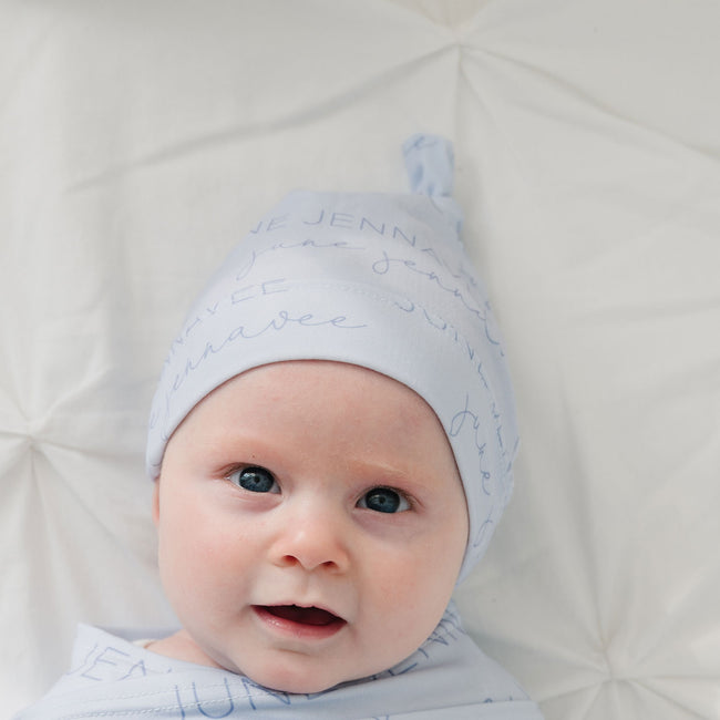 A close-up of a baby lying on a white textured blanket, wearing a light blue knit hat with blue cursive text and a matching swaddle.