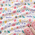 A close-up of a white fabric with a colorful watercolor-style print of flowers, leaves, ladybugs, bees, and the name 'Amelia' repeated in purple script. A pink plush butterfly toy is partially visible in the lower-right corner.