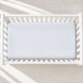 A light blue and white chevron-patterned fitted crib mattress inside a white wooden crib, photographed from a top-down angle on a light textured rug.