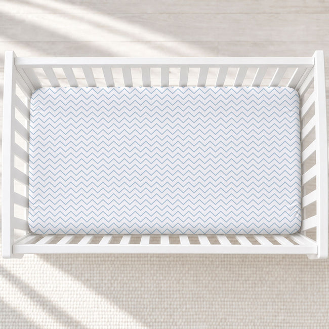 A light blue and white chevron-patterned fitted crib mattress inside a white wooden crib, photographed from a top-down angle on a light textured rug.