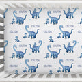 A crib fitted sheet featuring a repeating pattern of blue watercolor long-neck dinosaurs and the name 'COLTON' printed in blue on a white background, shown inside a white wooden crib.