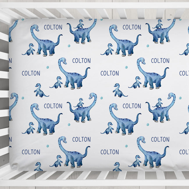 A crib fitted sheet featuring a repeating pattern of blue watercolor long-neck dinosaurs and the name 'COLTON' printed in blue on a white background, shown inside a white wooden crib.