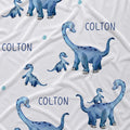White fabric with a repeating pattern of blue watercolor dinosaurs and the name 'COLTON' printed in dark blue text.