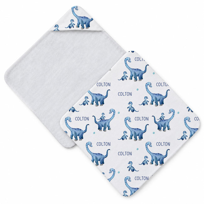A square baby towel shown from two angles on a white background. One side shows a soft white reverse, and the other shows a white front printed with blue dinosaurs and the name 'COLTON' repeated in blue text.