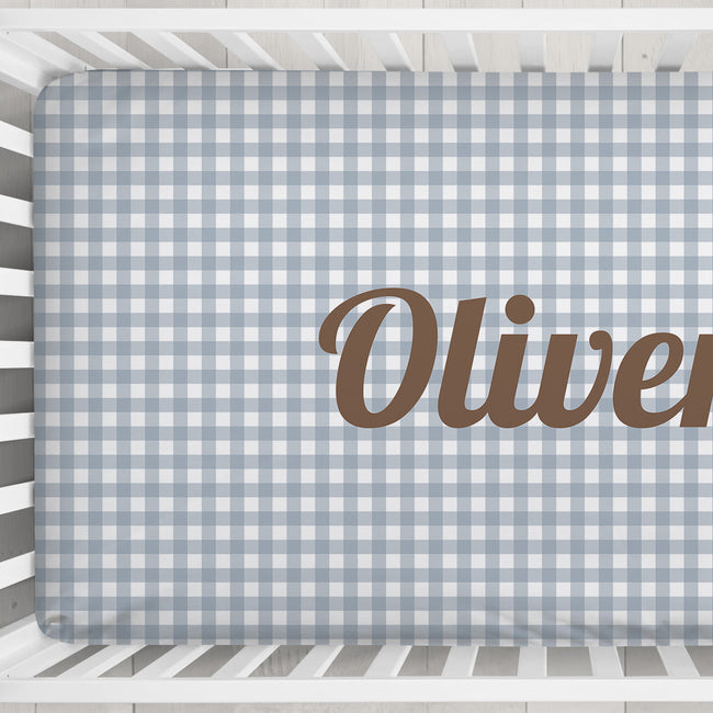 A fitted crib mattress with a light blue and white gingham check pattern and the name 'Oliver' printed in brown script across the right side, shown inside a white wooden crib.