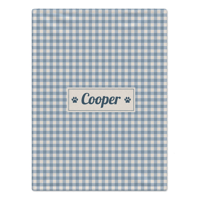 A rectangular personalized dog blanket with a light blue and white gingham check pattern, featuring a central cream label with the name 'Cooper' in navy blue script and navy paw prints on either side, shown on a plain white background.