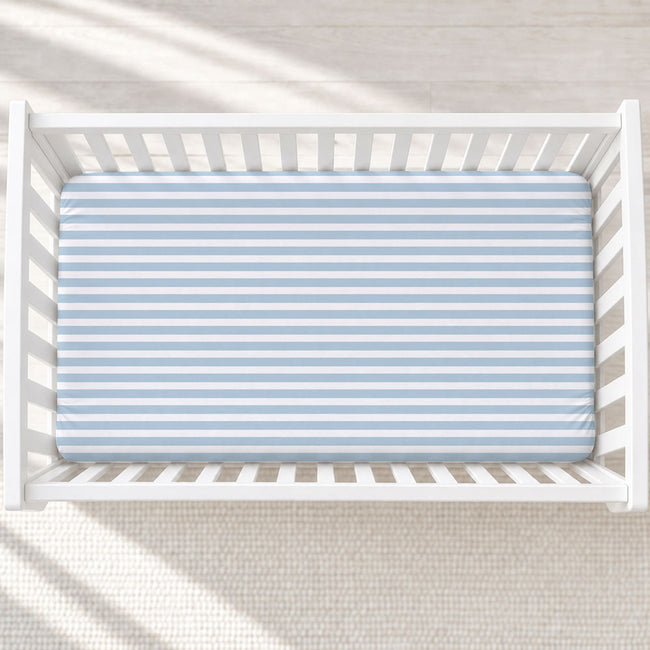 A top-down view of a white wooden crib with a light blue and white striped fitted sheet, set against a neutral cream textured rug with soft diagonal shadows.