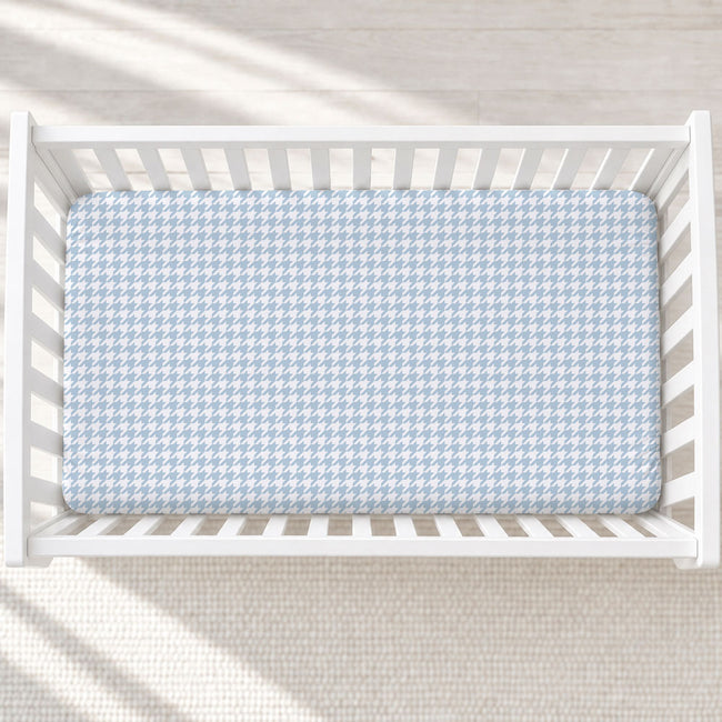A light blue and white houndstooth patterned mattress protector fitted inside a white wooden baby crib, shown from a top-down perspective on a light textured rug.