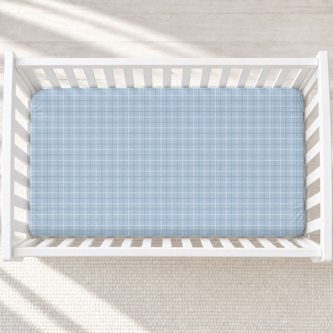A light blue and white plaid fitted crib mattress shown inside a white wooden baby crib, photographed from directly above on a light textured rug.