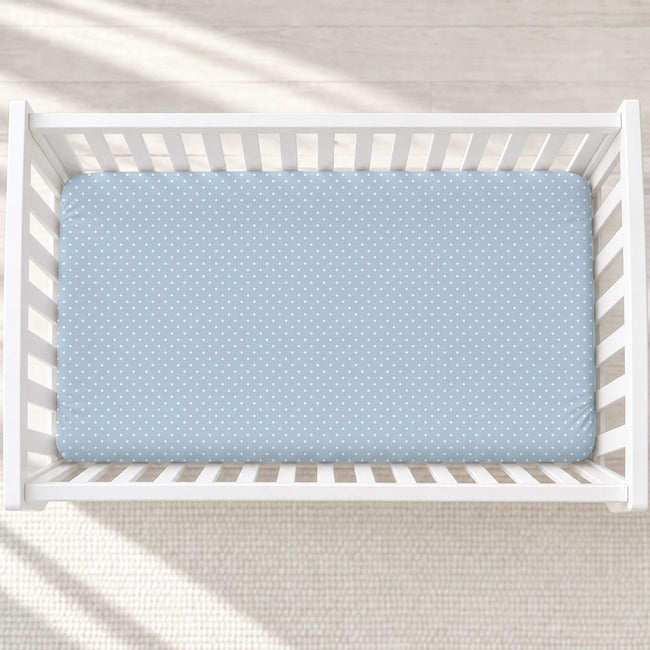A light blue fitted crib sheet with a white polka dot pattern, shown inside a white wooden baby crib on a light textured rug.