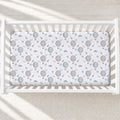 A fitted crib mattress with a white background and a repeating print of gray hot air balloons, small blue clouds, and tiny gold accents, shown inside a white wooden crib.