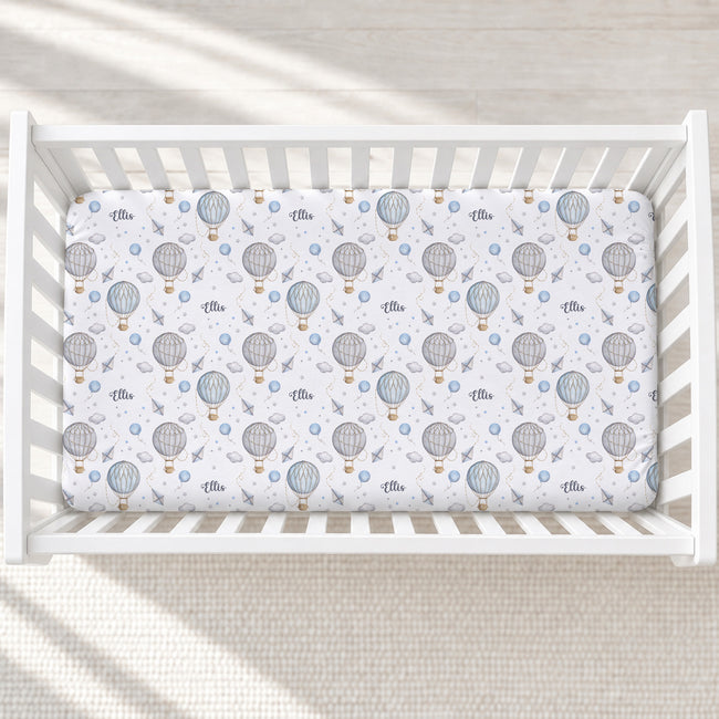 A fitted crib mattress with a white background and a repeating print of gray hot air balloons, small blue clouds, and tiny gold accents, shown inside a white wooden crib.
