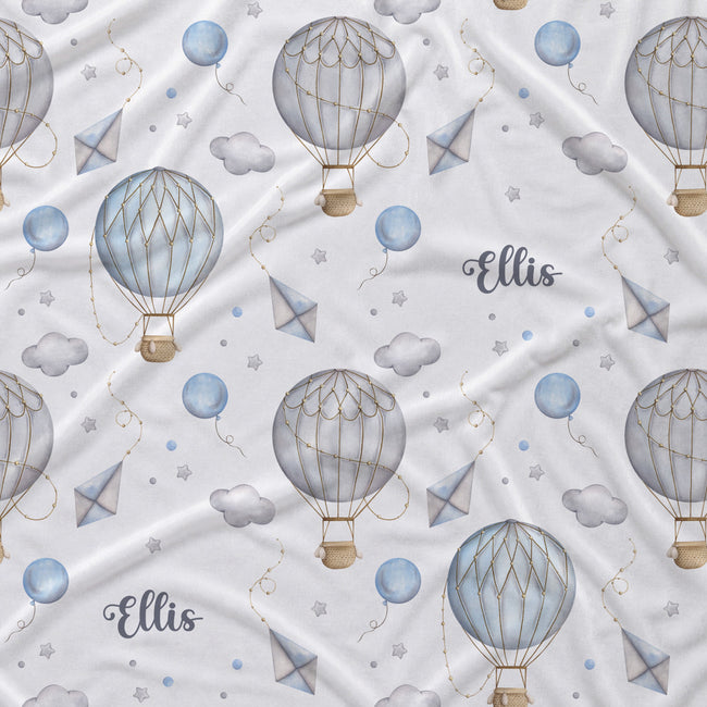 A white fabric with a repeating pattern of watercolor hot air balloons, clouds, stars, blue balloons, and the name 'Ellis' in gray script.