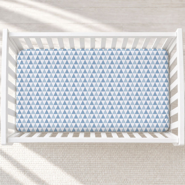 A top-down view of a white wooden crib with a fitted sheet featuring a repeating light blue and white triangle pattern, set against a light beige textured rug with soft window light shadows.