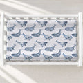 A baby changing table with a white changing pad cover featuring a repeating pattern of blue-gray whales, shown inside a white wooden crib on a light textured floor with soft window shadows.
