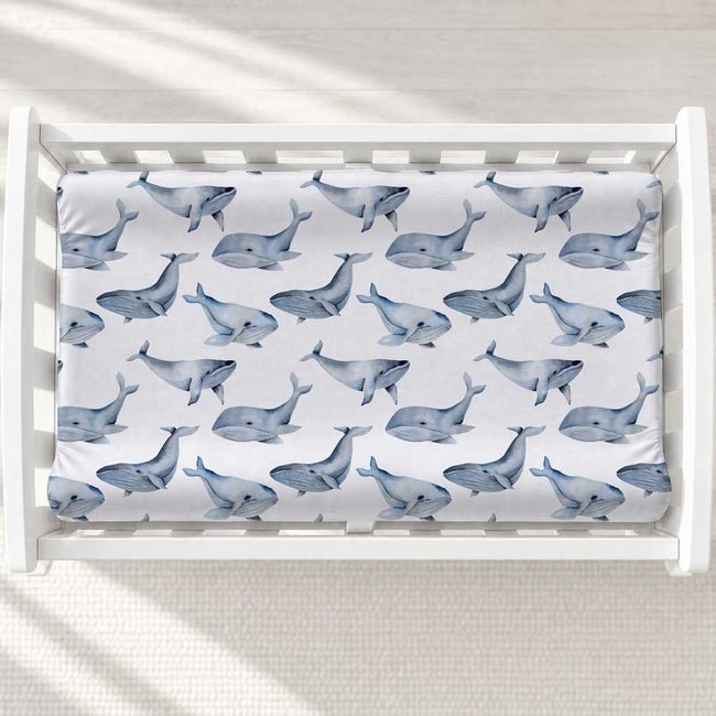 A baby changing table with a white changing pad cover featuring a repeating pattern of blue-gray whales, shown inside a white wooden crib on a light textured floor with soft window shadows.
