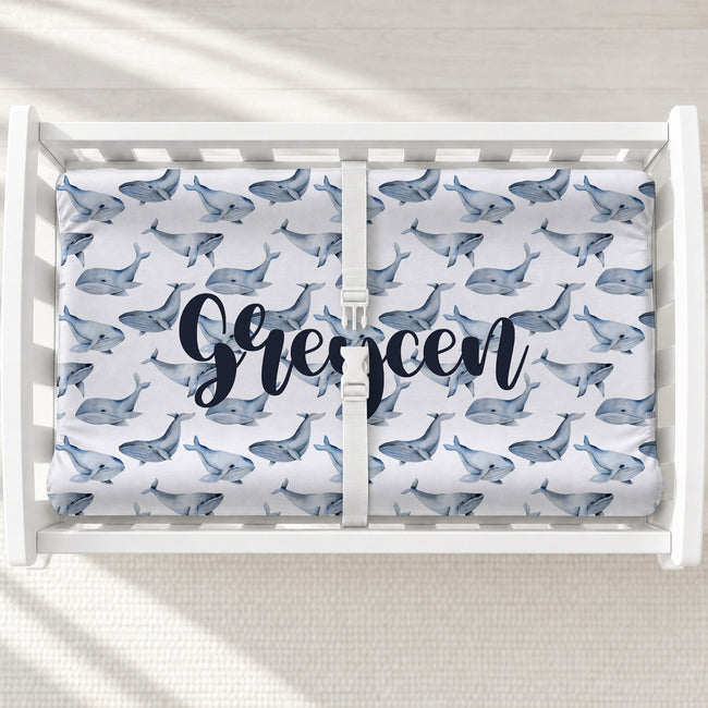 A personalized changing pad cover featuring a repeating blue whale print and the name 'Grecen' in large navy script across the center, shown from above in a bright nursery.