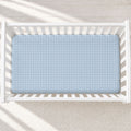 A light blue fitted crib sheet with a white windowpane pattern, shown inside a white wooden baby crib on a light textured rug.