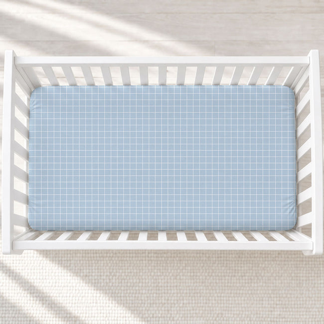A light blue fitted crib sheet with a white windowpane pattern, shown inside a white wooden baby crib on a light textured rug.