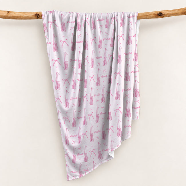 Personalized swaddle blanket hanging on a wooden rail with pink watercolor ballet slippers, bows and custom name Elizabeth.