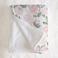 A folded baby blanket with a soft white fleece reverse side and a printed side featuring pink roses and green leaves on a white background, styled on a white faux fur surface.