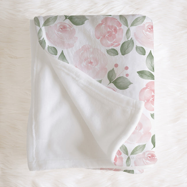 A folded baby blanket with a soft white fleece reverse side and a printed side featuring pink roses and green leaves on a white background, styled on a white faux fur surface.