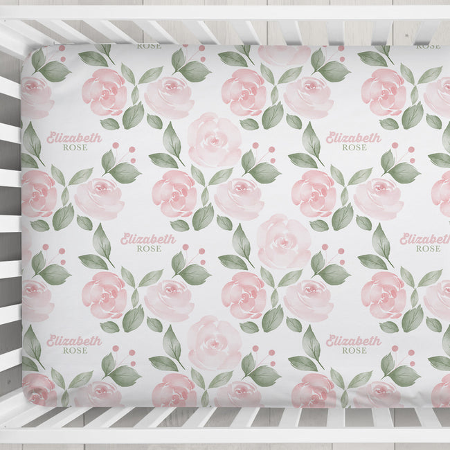 Close-up of Blush Rose Garden crib sheet with pink watercolor roses, sage leaves, and Elizabeth Rose lettering in a white crib.