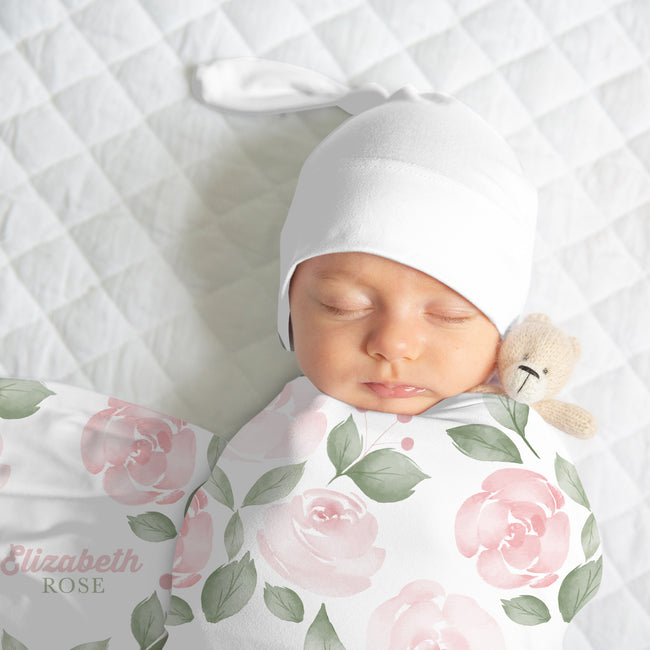 Newborn wearing a white knotted hat, wrapped in a Blush Rose Garden swaddle with pink roses, sage leaves, and Elizabeth Rose lettering.