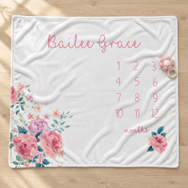 A personalized baby blanket laid flat on a light wood floor. The white blanket features pink script text reading 'Bailee Grace' at the top, a grid of numbers 1 through 12 for months, and pink floral prints with green leaves in the bottom corners.
