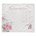 A white personalized baby blanket featuring pink script text reading 'Bailee Grace,' a pink numbered monthly calendar grid from 1 to 12, and pink floral illustrations with green leaves in the corners.