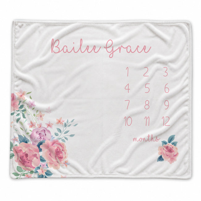 A white personalized baby blanket featuring pink script text reading 'Bailee Grace,' a pink numbered monthly calendar grid from 1 to 12, and pink floral illustrations with green leaves in the corners.