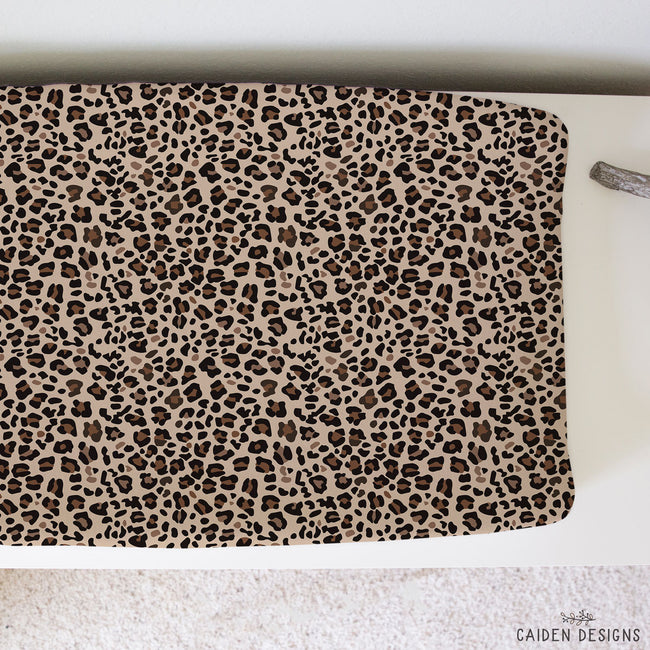 A baby changing pads cover featuring a classic leopard print pattern in beige, brown, and black. It is laid flat on a white surface with a textured white rug underneath.