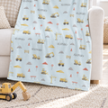 A light blue baby blanket with a construction-themed print of yellow excavators, cranes, and the name 'ALONSO' repeated, draped over a white sofa in a bright nursery setting.