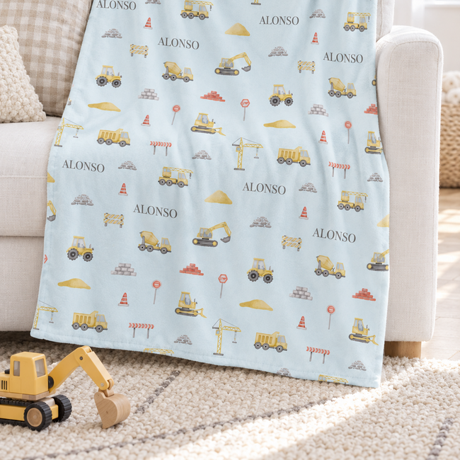 A light blue baby blanket with a construction-themed print of yellow excavators, cranes, and the name 'ALONSO' repeated, draped over a white sofa in a bright nursery setting.