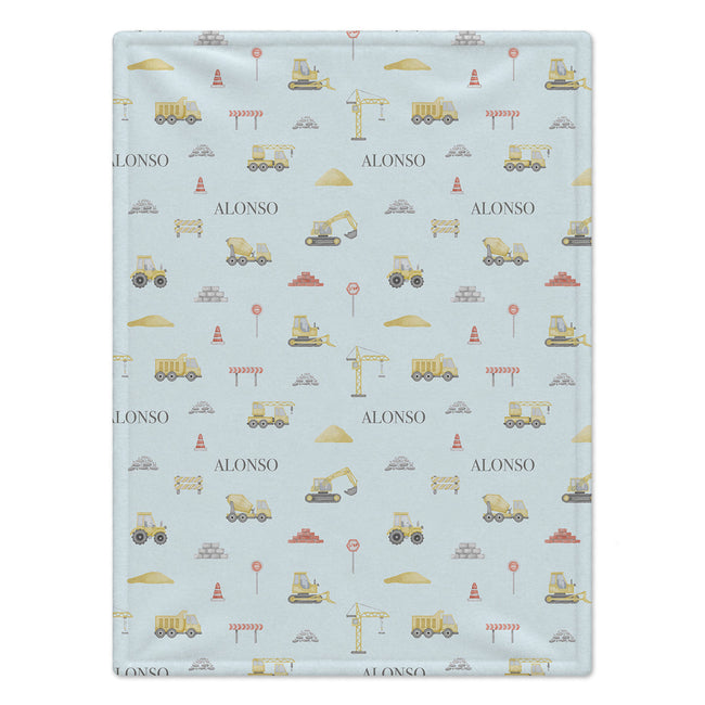 A personalized baby blanket with a light blue background and a construction-themed pattern featuring yellow excavators, dump trucks, cranes, and the name 'ALONSO' printed repeatedly.