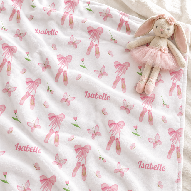 A white baby blanket with a pink ballet theme print, featuring pink bows, ballerina shoes, butterflies, and the name 'Isabelle' repeated. A plush bunny doll wearing a pink tutu is resting on top of the blanket.