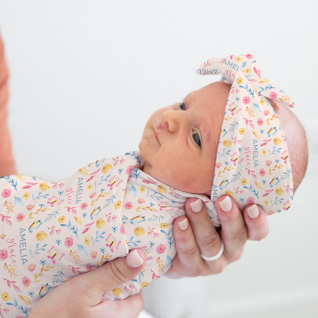 A baby swaddled in a pink floral fabric blanket and wearing a matching pink floral headband with the name 'AMELIA' printed on it, held by an adult's hands against a bright white background.