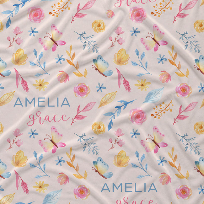 A close-up of a fabric surface with a repeating watercolor floral and butterfly print on a warm off-white background. The pattern includes pink and yellow flowers, blue and yellow leaves, and pink and blue butterflies.