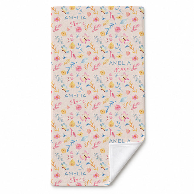 A personalized beach towel with a pastel floral and bird pattern on a cream background, featuring the names 'AMELIA' and 'Grace' printed repeatedly. The bottom corner is folded over to show the white reverse side.