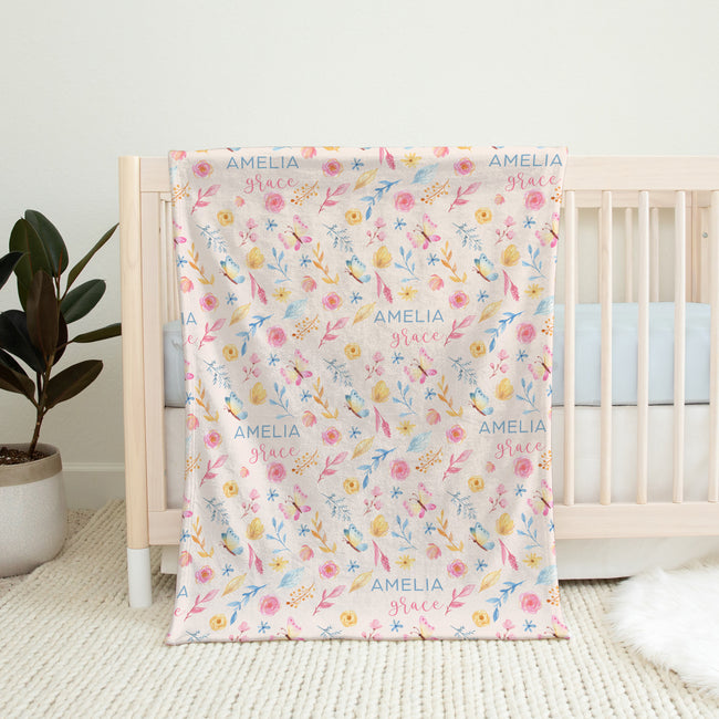 A personalized baby blanket with a floral pattern and the name 'AMELIA grace' printed repeatedly, draped over a light wood crib in a nursery setting.