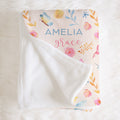 A folded baby blanket with a white plush fleece backing and a printed floral pattern on the top side. The blanket is personalized with the names 'AMELIA' and 'grace' in blue and pink lettering.