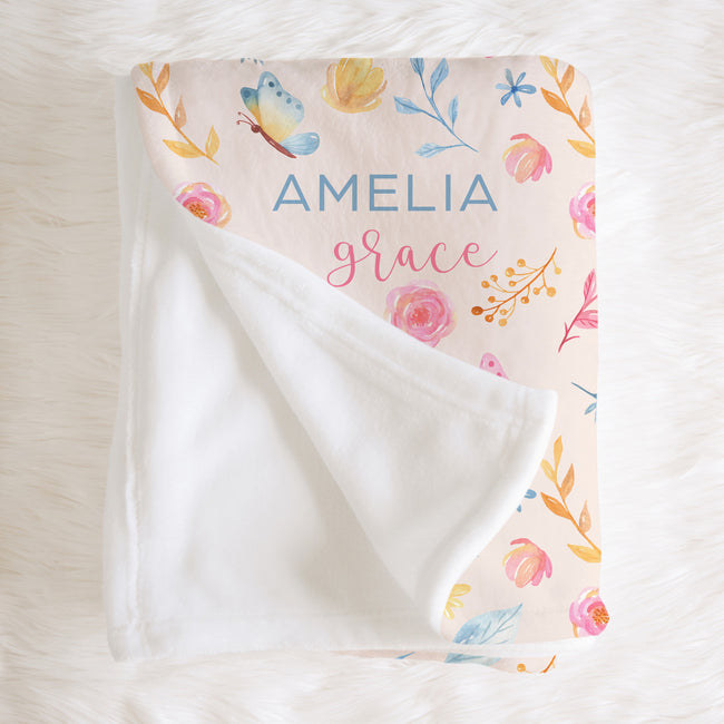 A folded baby blanket with a white plush fleece backing and a printed floral pattern on the top side. The blanket is personalized with the names 'AMELIA' and 'grace' in blue and pink lettering.