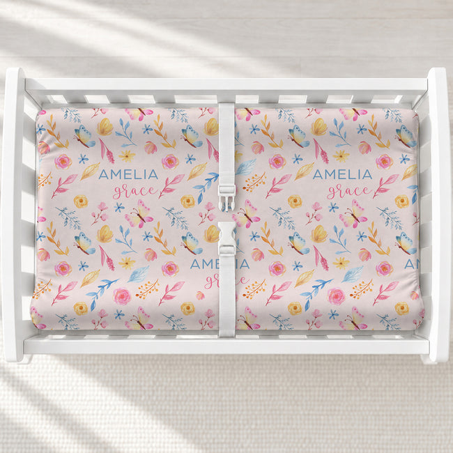 A white changing table with a personalized changing pad cover featuring a pastel floral and butterfly pattern. The fabric are personalized with the names 'AMELIA' and 'grace' in blue script.