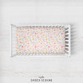 A rectangular crib mattress fitted with a pastel floral print sheet featuring the names 'Amelia Grace' repeated in blue script, displayed in a white wooden crib on a light whitewashed wood background. The brand name 'CAIDEN DESIGNS' is printed at the bottom.