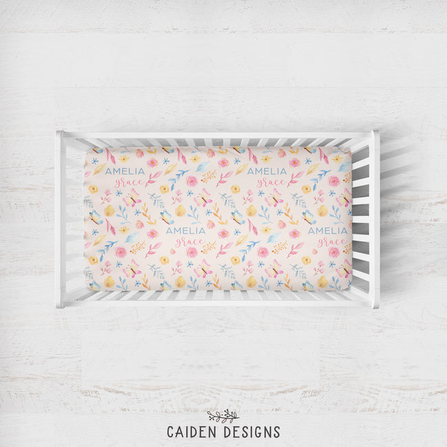 A rectangular crib mattress fitted with a pastel floral print sheet featuring the names 'Amelia Grace' repeated in blue script, displayed in a white wooden crib on a light whitewashed wood background. The brand name 'CAIDEN DESIGNS' is printed at the bottom.