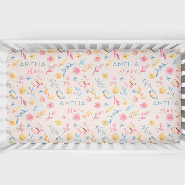 A crib mattress fitted sheet with a pastel watercolor floral pattern and the personalized text 'AMELIA grace' printed repeatedly, shown inside a white wooden crib.