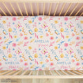 A crib mattress fitted sheet with a colorful watercolor floral and butterfly pattern, personalized with the name 'AMELIA grace' in blue and pink script. The sheet is shown inside a light wood crib, viewed from above.