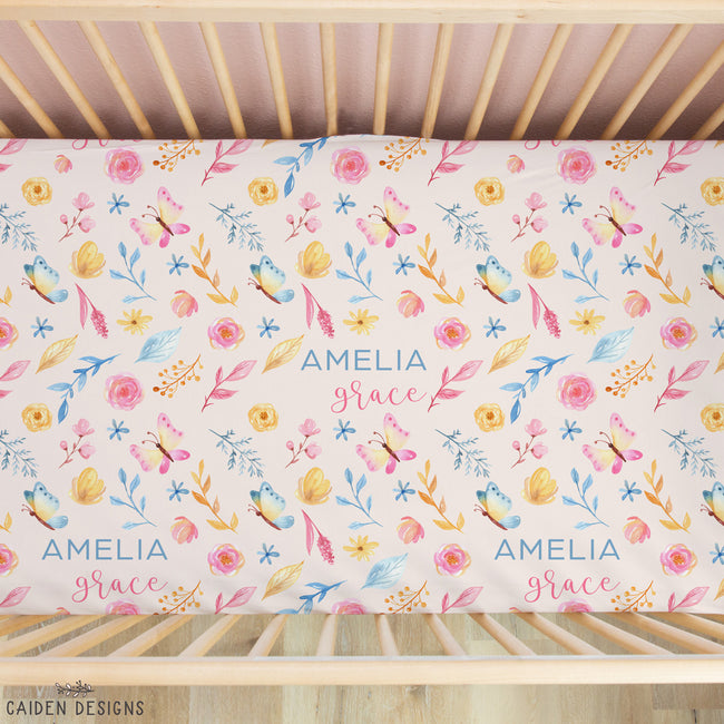 A crib mattress fitted sheet with a colorful watercolor floral and butterfly pattern, personalized with the name 'AMELIA grace' in blue and pink script. The sheet is shown inside a light wood crib, viewed from above.
