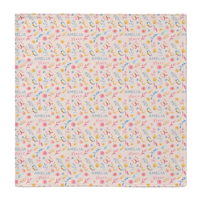 A square personalized baby swaddle blanket with a delicate floral and bird pattern on a light pink background, featuring the name 'AMELIA' printed repeatedly in blue.